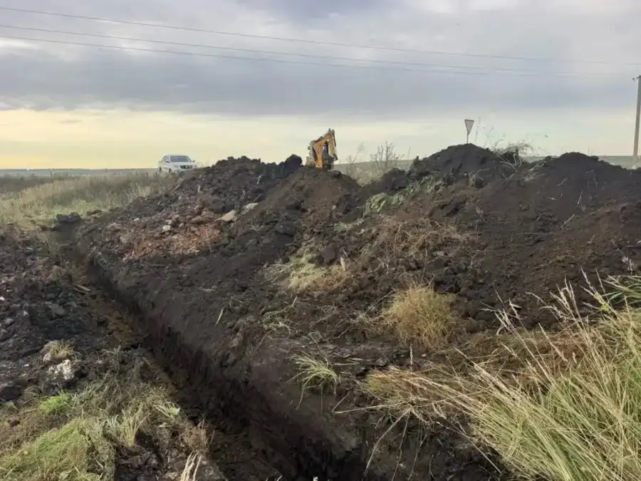 Глубокая траншея и отвалы грунта в поле, вдали виден экскаватор.
