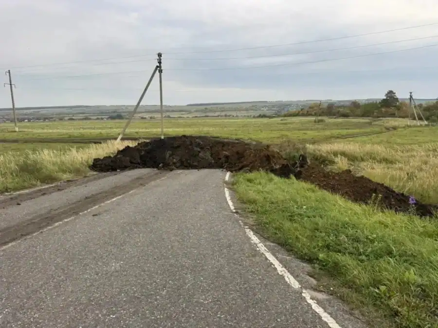 Насыпь грунта перекрывает узкую асфальтированную дорогу среди полей.