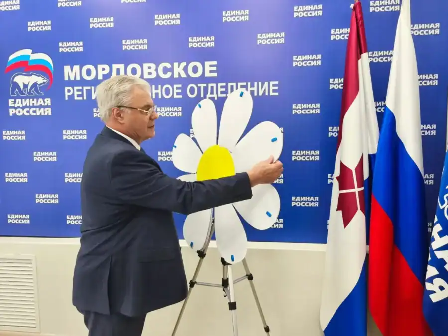 Мужчина снимает лепесток с ромашки на стойке рядом с флагами.