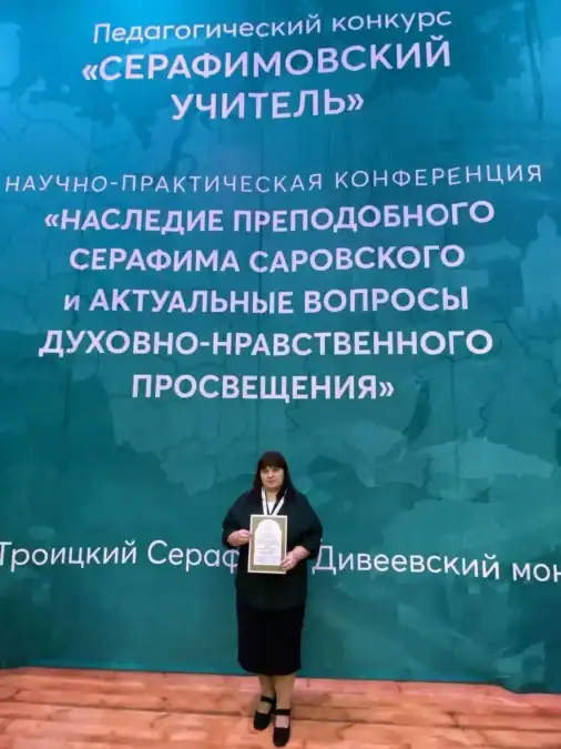Женщина с дипломом стоит у баннера педагогического конкурса.