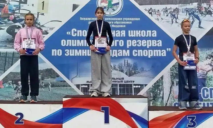 Три юные спортсменки с медалями и грамотами стоят на пьедестале.