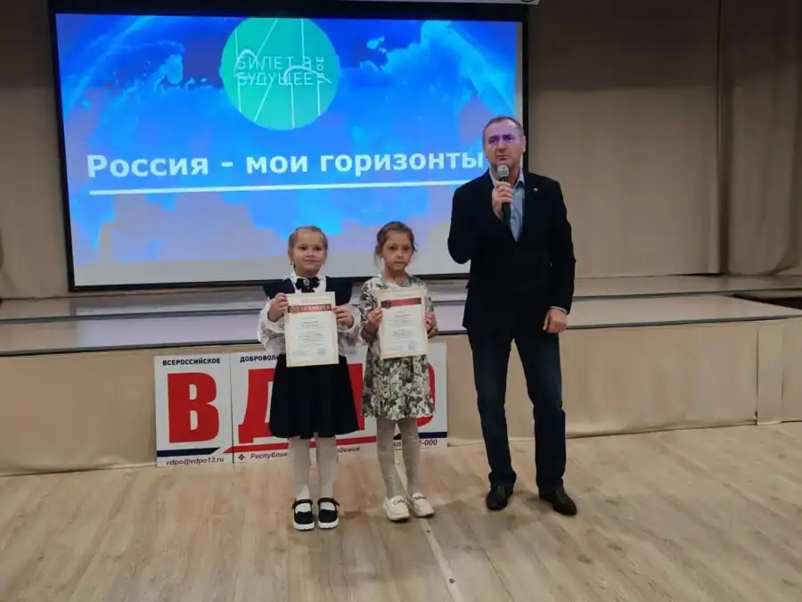 Две девочки с дипломами стоят на сцене рядом с мужчиной с микрофоном.