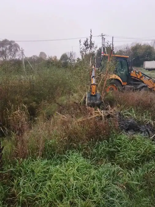 Экскаватор очищает заросшую канаву возле травы и кустарника.