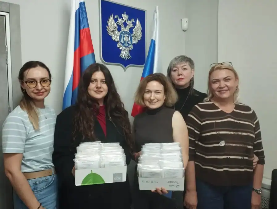 Пять сотрудниц держат коробки с упакованными комплектами на фоне российских флагов.