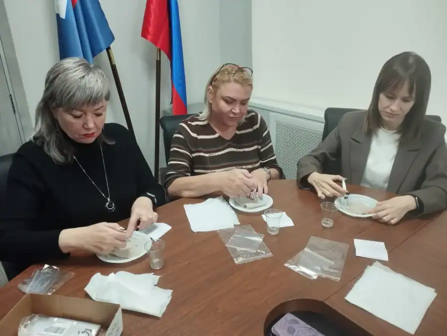 Три женщины за столом собирают небольшие комплекты на пластиковых тарелках.