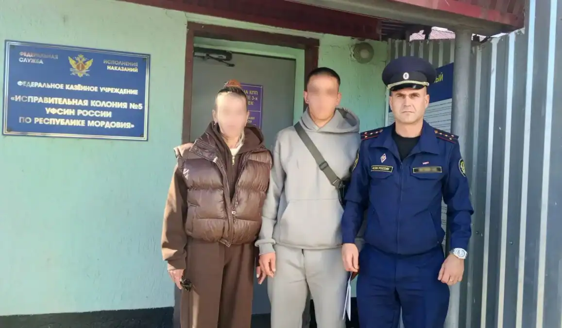 Трое мужчин стоят у входа в исправительную колонию рядом с ведомственной вывеской.