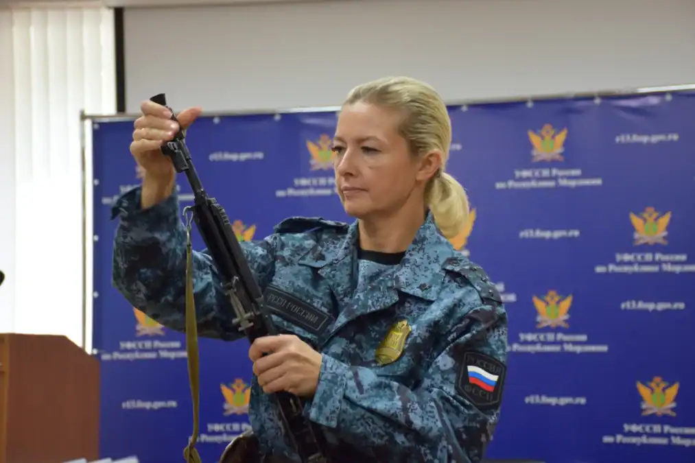 Сотрудница в камуфляжной форме держит автомат перед служебным баннером.