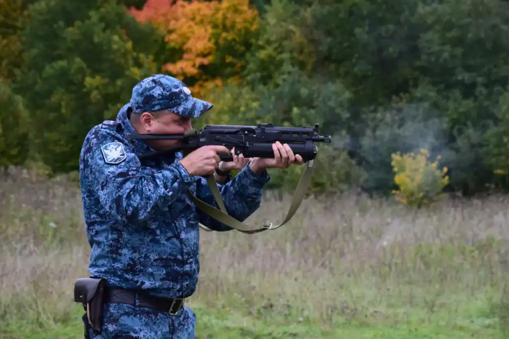 Сотрудник в камуфляжной форме стреляет из автомата на открытой площадке.