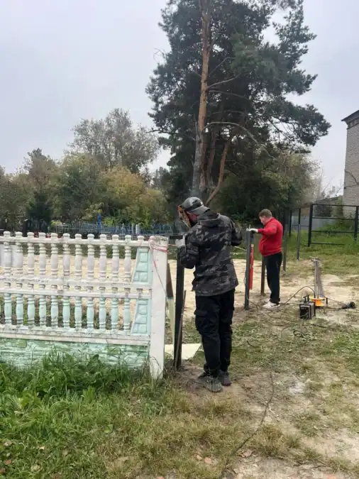 Двое мужчин устанавливают металлические стойки возле кладбищенской ограды.