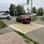 Бордовый седан у перехода, рядом видны служебный автомобиль и лежащий самокат.