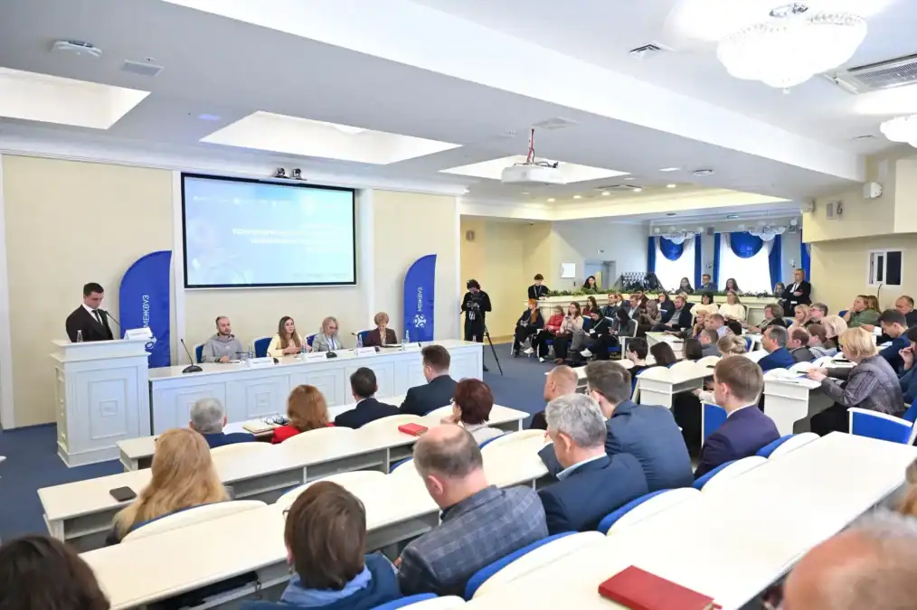 Выступающий у трибуны обращается к участникам в заполненном конференц-зале.