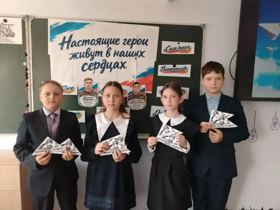 Четверо школьников держат письма-треугольники у стенда о героях.