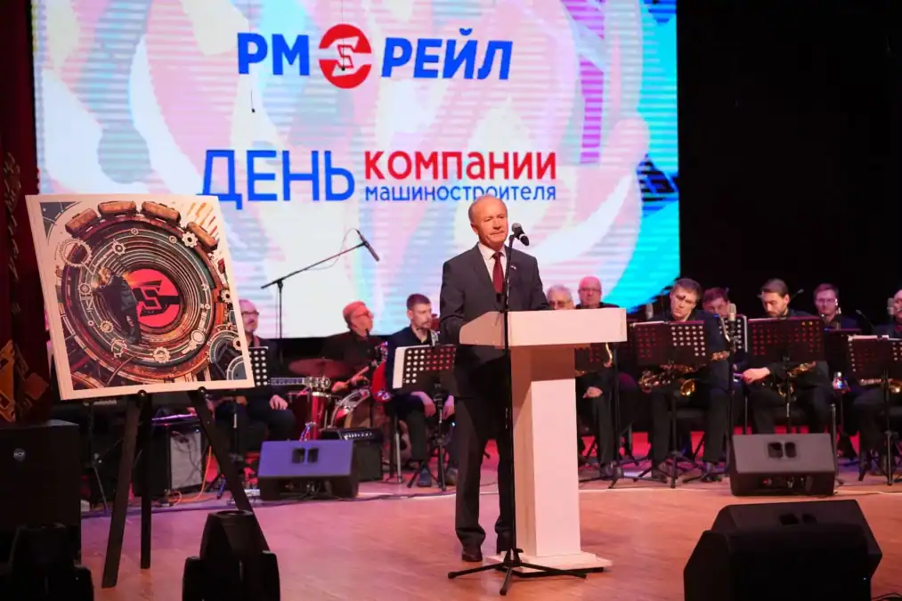 Мужчина выступает с трибуны на сцене перед оркестром.