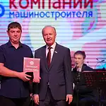 Двое мужчин на сцене держат красную награду перед оркестром.