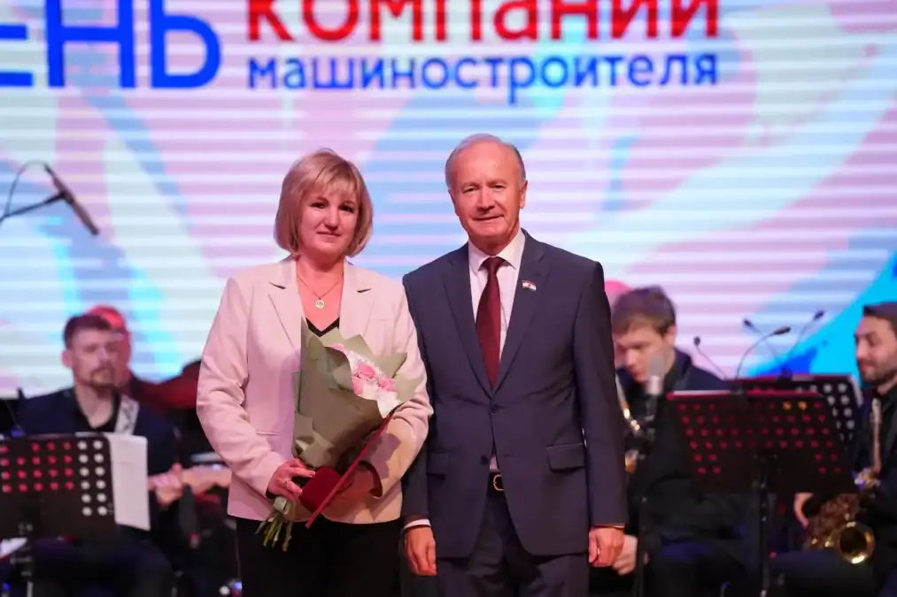 Женщина с букетом стоит на сцене рядом с мужчиной в костюме.