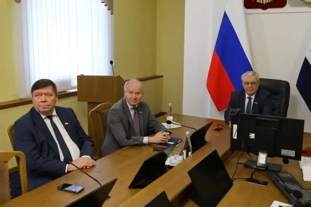 Трое мужчин сидят за столом в кабинете с российским флагом.