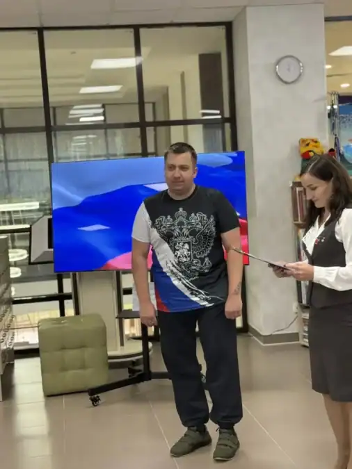 Мужчина стоит рядом с женщиной, читающей лист, на фоне экрана с российским флагом.