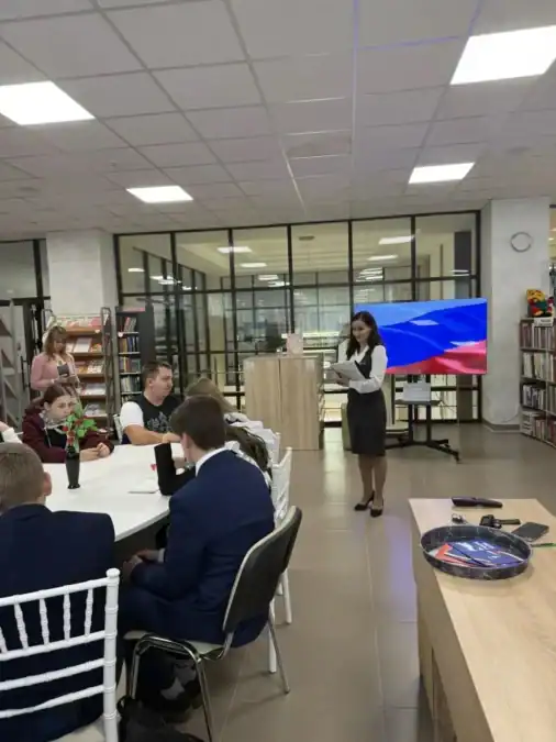 Подростки сидят за столом и слушают выступающую женщину в помещении с книжными стеллажами.