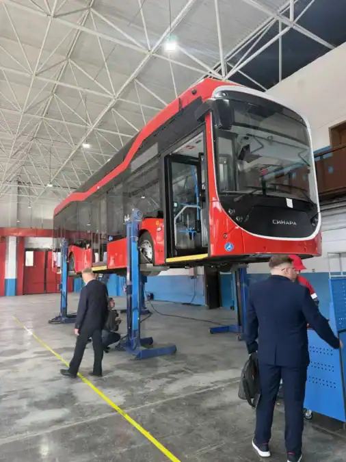 Красный электробус поднят на подъёмниках в производственном цехе.