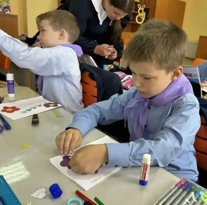 Школьник приклеивает бумажный цветок к открытке, рядом работают другие дети.