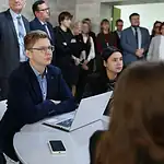 Мужчина с ноутбуком сидит за столом среди студентов и сотрудников.