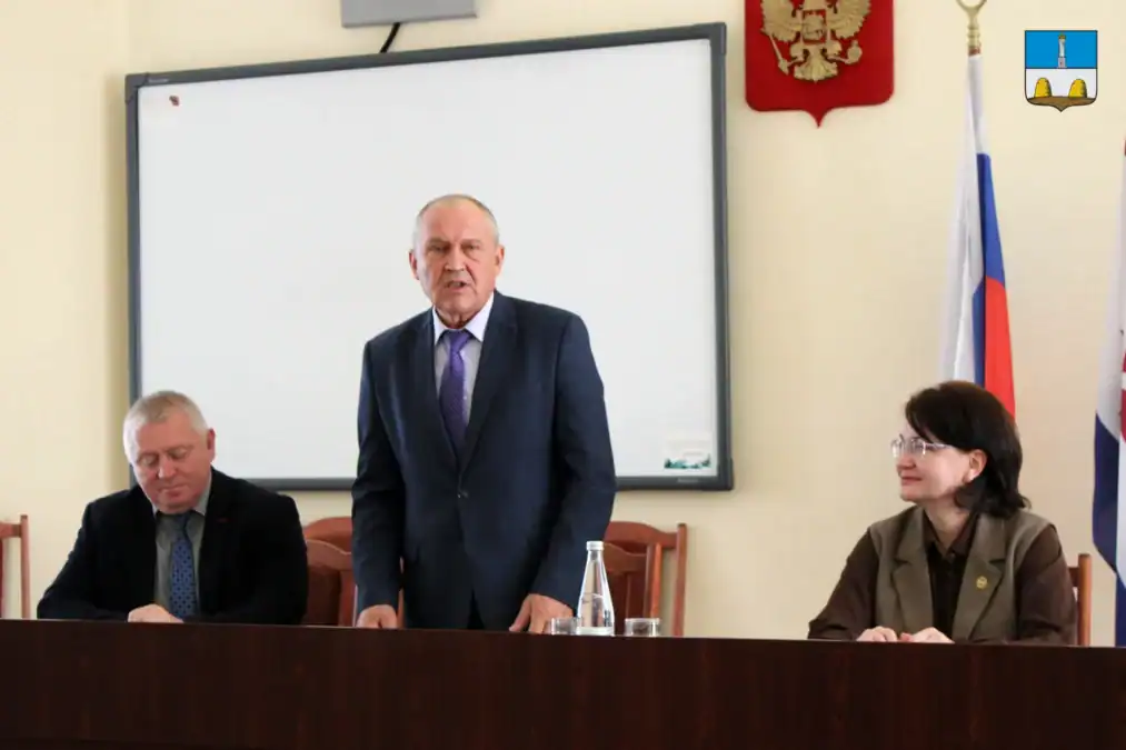 Мужчина выступает за столом президиума рядом с двумя участниками заседания.