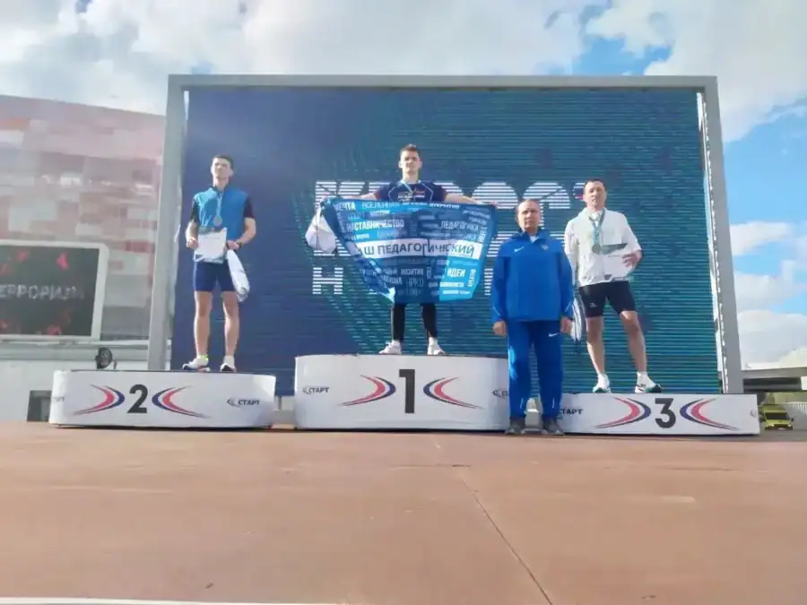 Спортсмены с дипломами стоят на трёх ступенях пьедестала перед большим экраном.