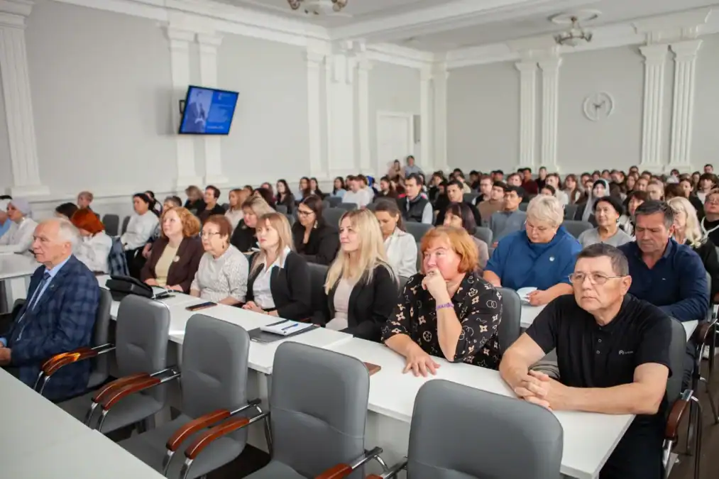 Участники конференции сидят в заполненном зале за столами.