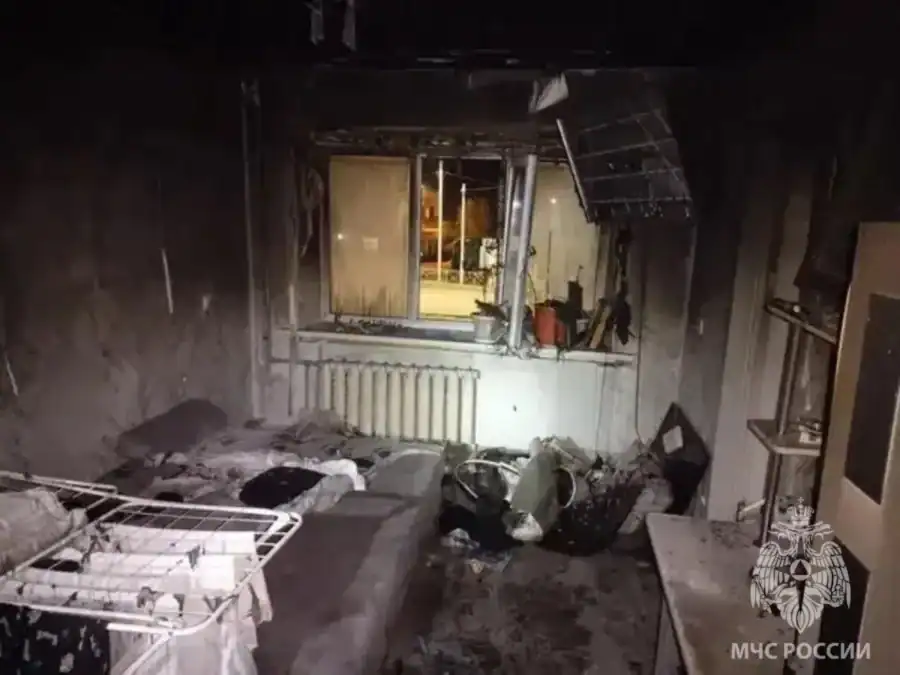 В выгоревшей комнате видны закопчённые стены, повреждённая мебель и вещи на полу.
