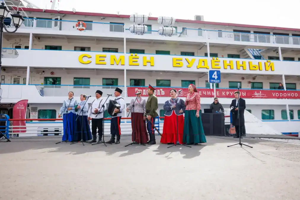 Артисты в народных костюмах выступают с микрофонами перед теплоходом.