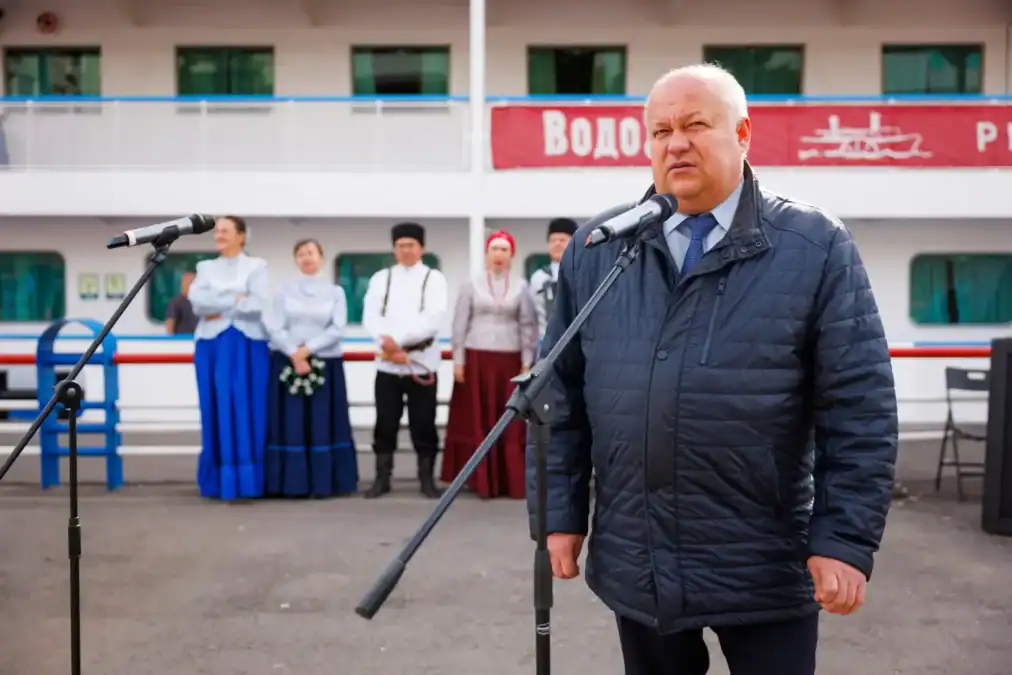 Мужчина выступает у микрофона, позади стоят артисты в народных костюмах.