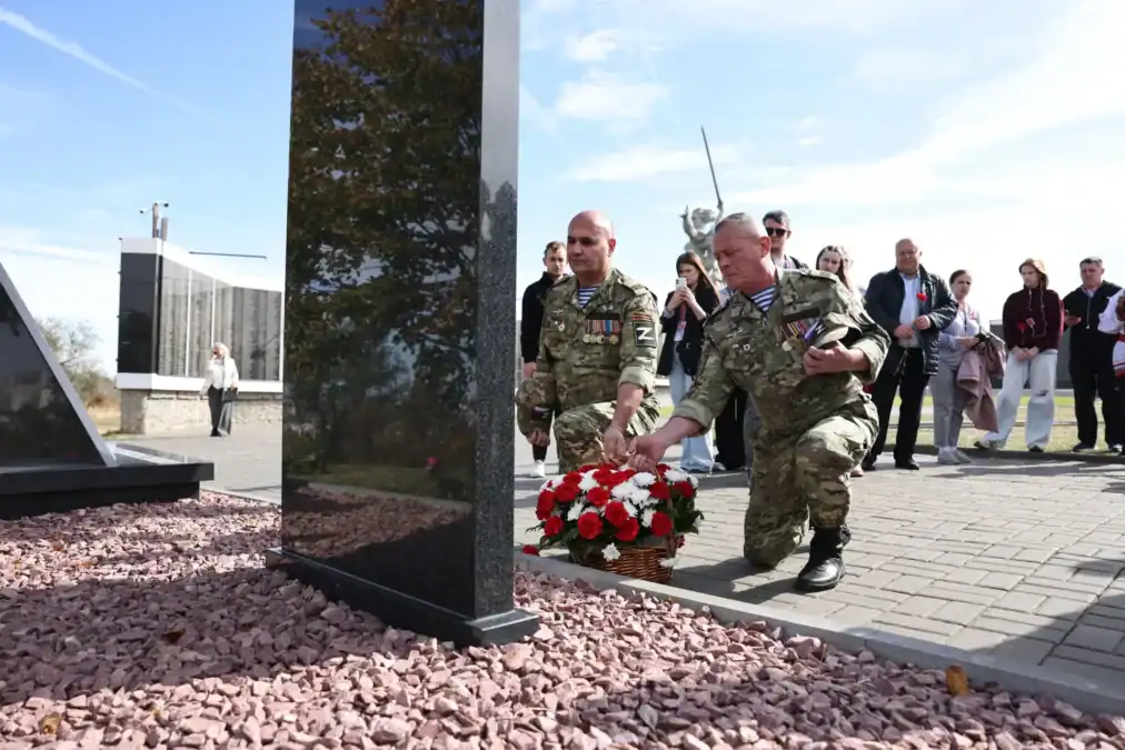 Двое мужчин в камуфляже возлагают корзину красных гвоздик к памятному знаку.