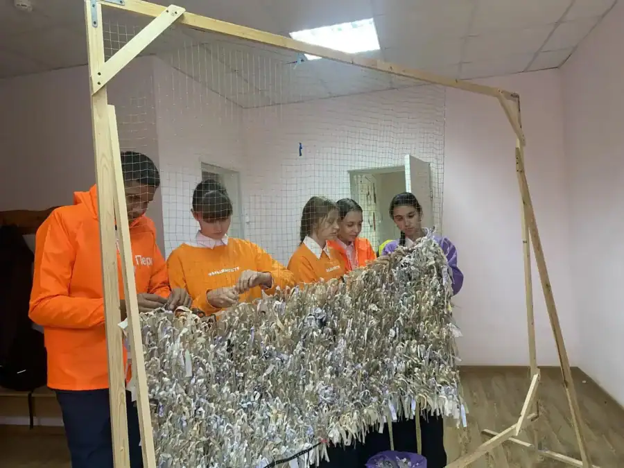 Волонтёры вплетают тканевые лоскуты в большую маскировочную сеть на деревянной раме.