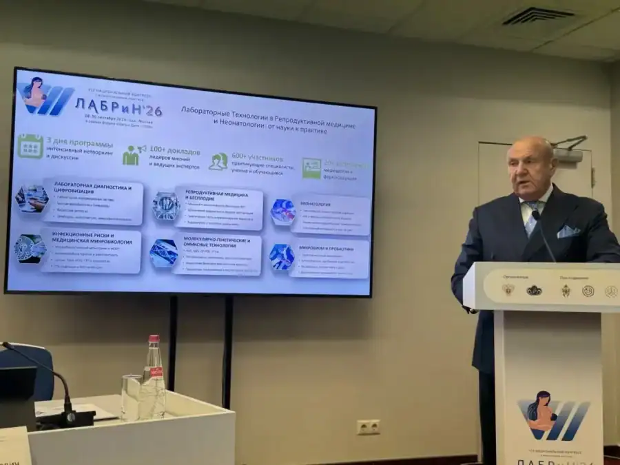 Мужчина выступает с трибуны рядом с экраном презентации форума.