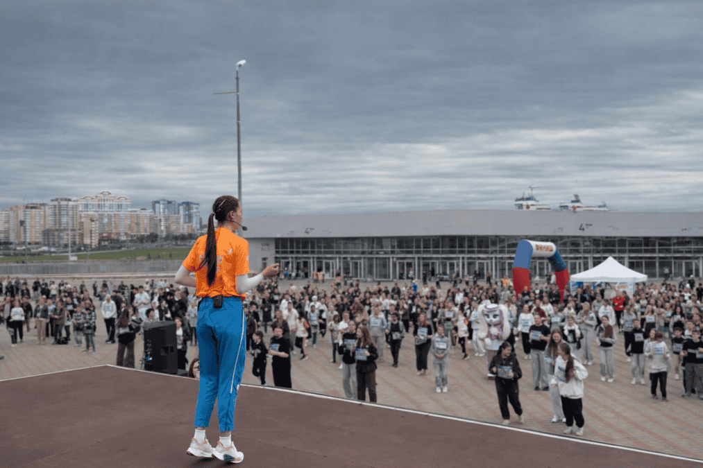 Инструктор в оранжевой футболке проводит зарядку перед участниками на площадке у стадиона.