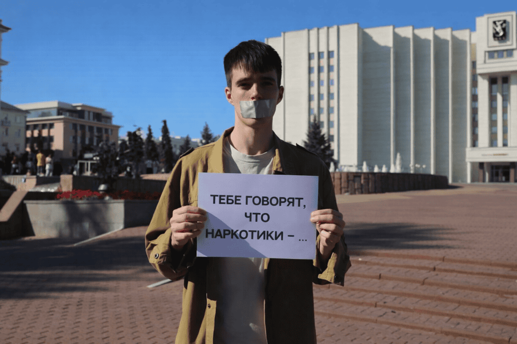 Молодой человек с заклеенным ртом держит плакат с предупреждением о наркотиках.