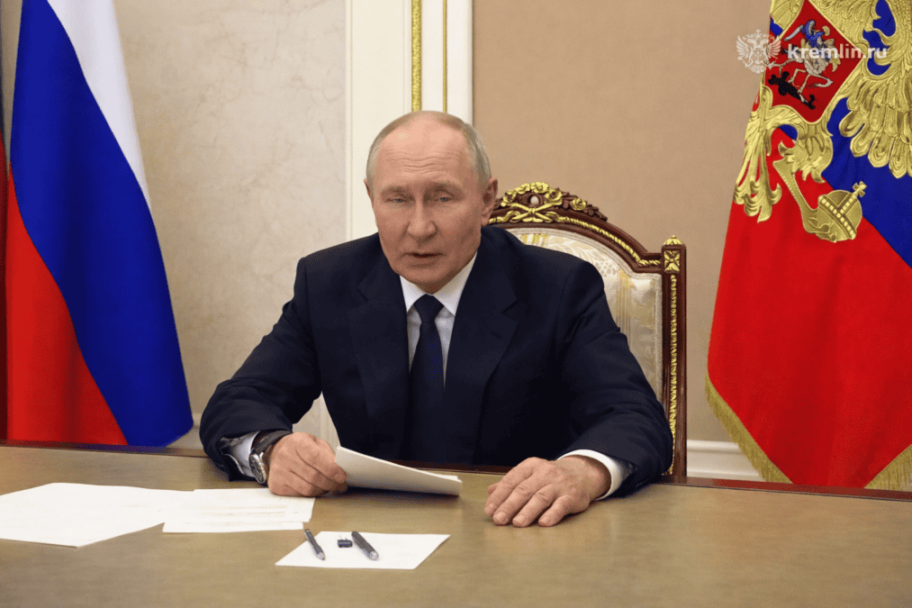Президент России сидит за столом с документом на фоне российских флагов.