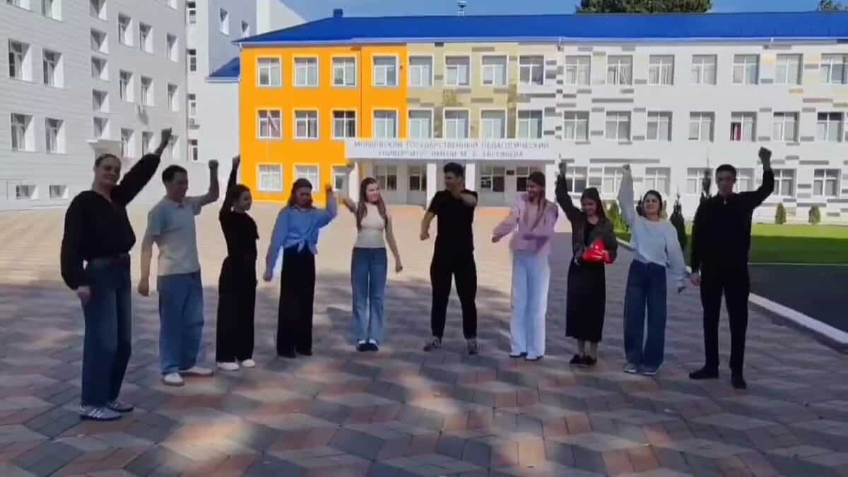 Группа студентов с поднятыми руками стоит перед зданием университета.