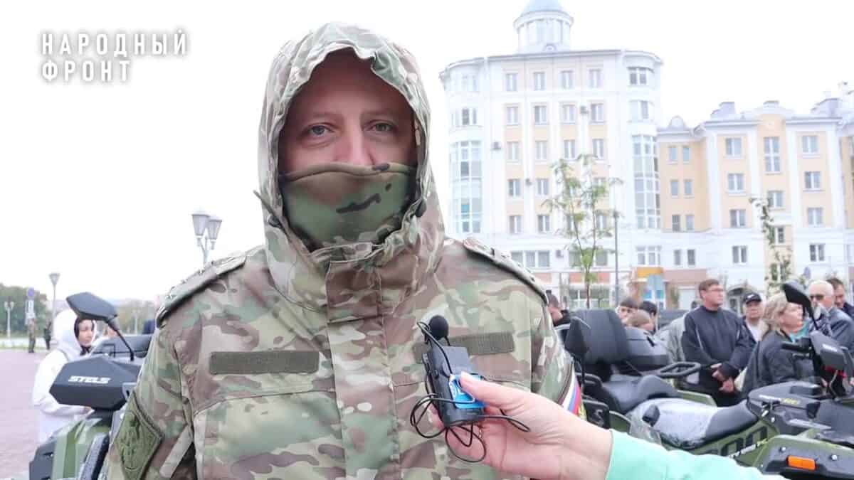 Военнослужащий в камуфляже даёт интервью на фоне квадроциклов и людей.