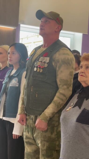 Мужчина с медалями стоит рядом с молодыми участницами мероприятия.