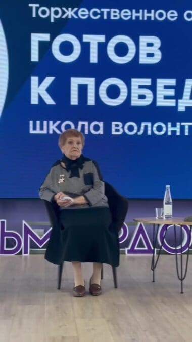 Женщина сидит на сцене перед экраном с названием школы.