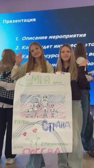 Две девушки держат плакат с рисунками и патриотическими надписями.