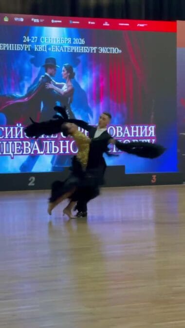 Танцевальная пара в движении выступает перед экраном соревнований.