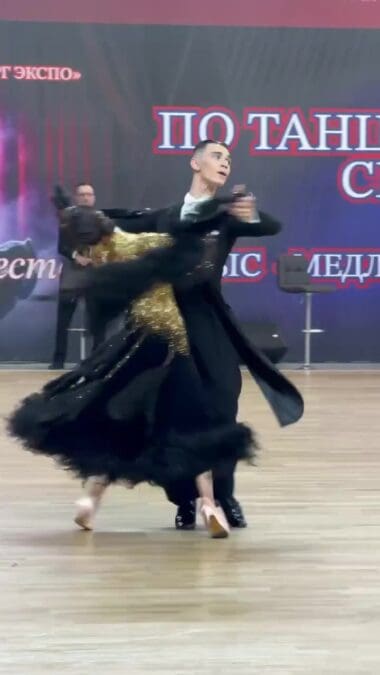 Танцоры в чёрно-золотых костюмах исполняют вращение на паркете.