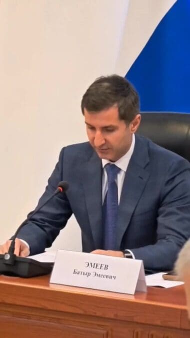 Мужчина в костюме выступает за столом с микрофоном на заседании.