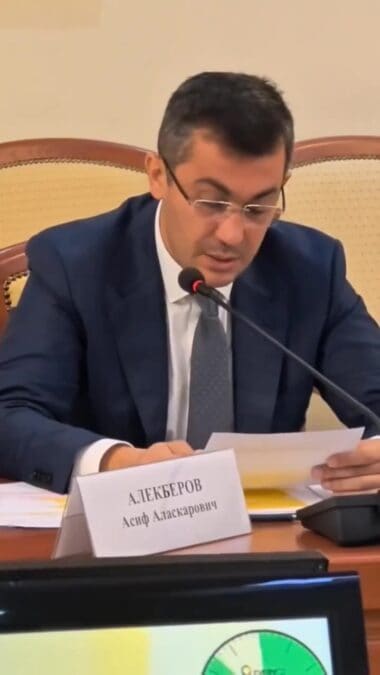 Мужчина в очках читает документ за столом с включённым микрофоном.