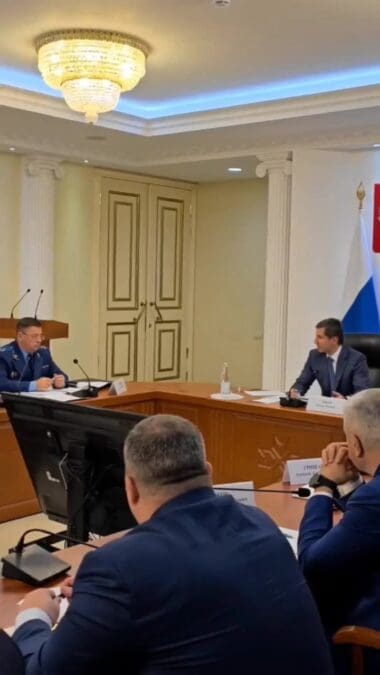 Участники заседания сидят за длинным столом в зале с российским флагом.