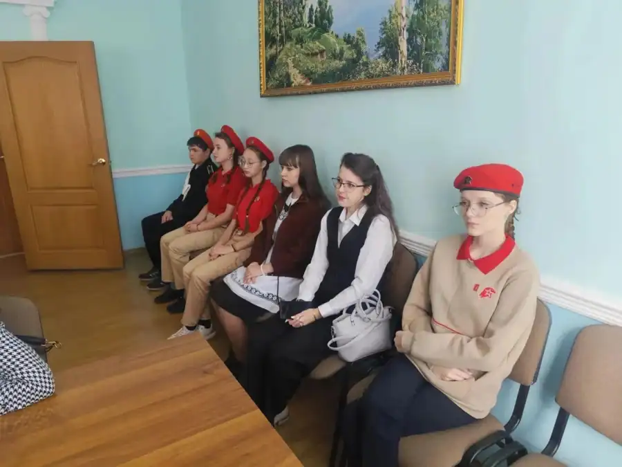 Шесть молодых участников сидят у стены в помещении администрации.