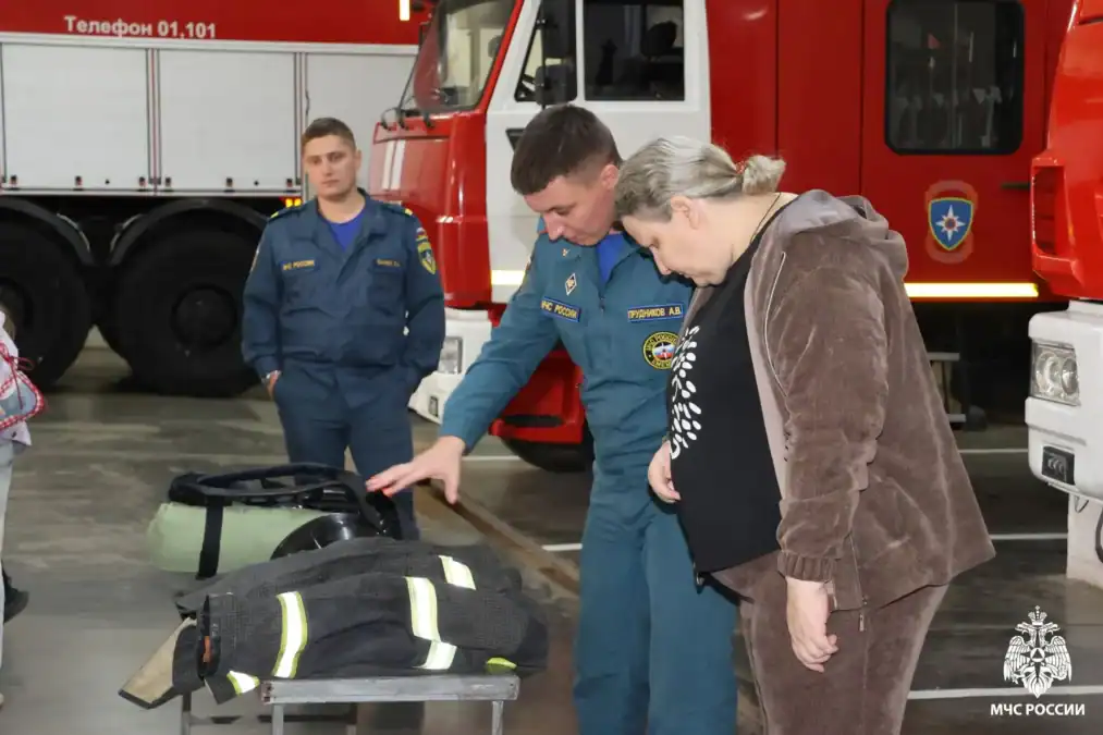 Сотрудник МЧС показывает женщине элементы пожарного снаряжения у пожарной машины.