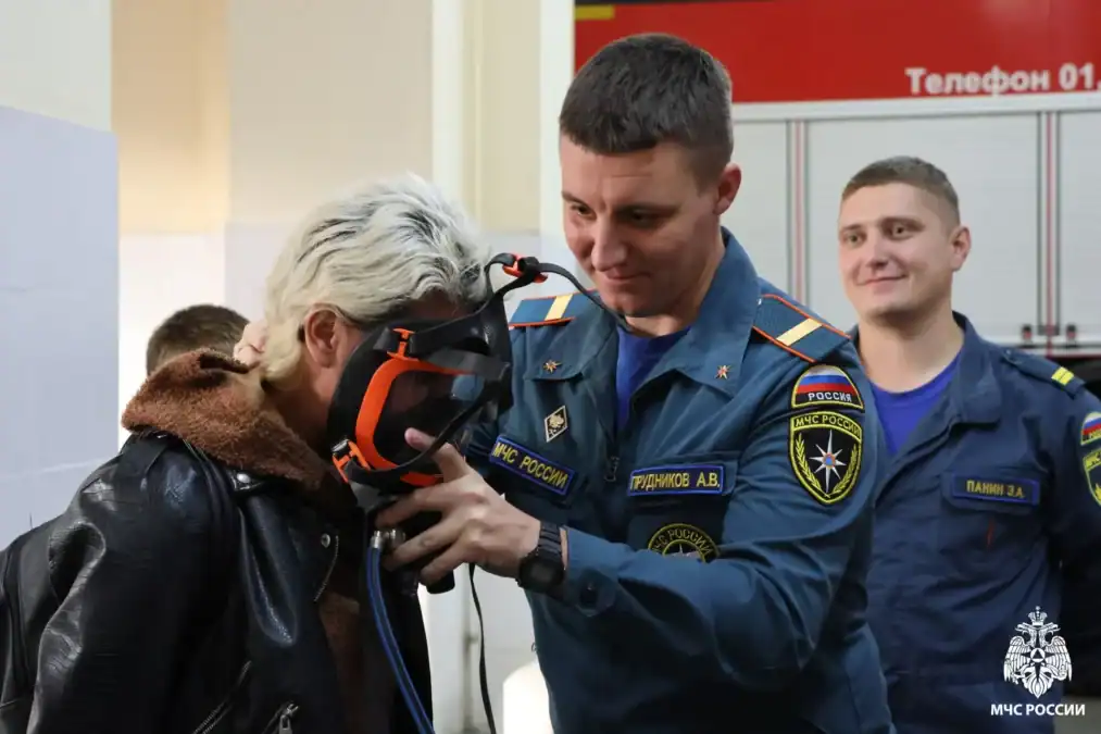 Сотрудник МЧС помогает женщине примерить дыхательный аппарат в пожарной части.